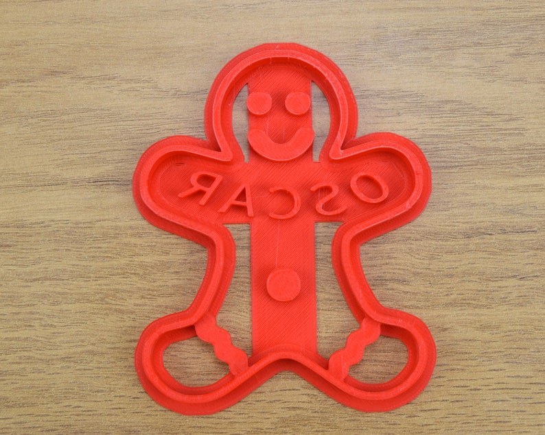 Puede incluir: Cortador de galletas de jengibre de pl&aacute;stico rojo en forma de hombre con la palabra "OSCAR" escrita en la parte delantera.