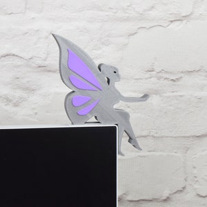 K&ouml;nnte beinhalten: Eine graue Feenfigur mit lila Fl&uuml;geln sitzt auf einem schwarzen Computermonitor. Die Fee tr&auml;gt ein langes, flie&szlig;endes Kleid und h&auml;lt ein kleines Objekt in der Hand.