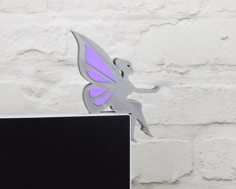 K&ouml;nnte beinhalten: Eine graue Feenfigur mit lila Fl&uuml;geln sitzt auf einem schwarzen Computermonitor. Die Fee tr&auml;gt ein langes, flie&szlig;endes Kleid und h&auml;lt ein kleines Objekt in der Hand.
