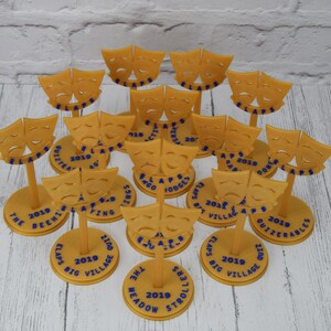 May include: A collection of 14 gold-colored 3D printed trophies with a theatrical mask design. Each trophy has a round base with blue text that includes the year 2019 and the name of a play or event.