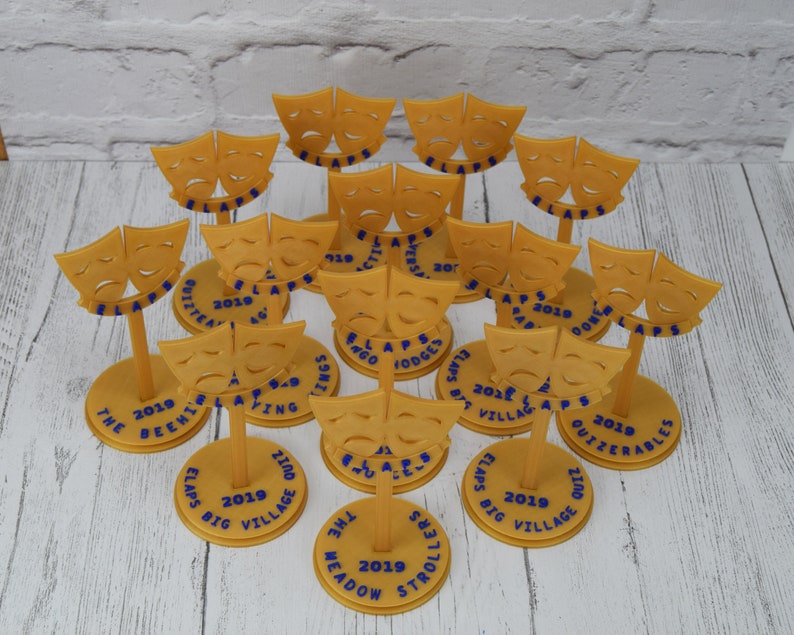 May include: A collection of 14 gold-colored 3D printed trophies with a theatrical mask design. Each trophy has a round base with blue text that includes the year 2019 and the name of a play or event.
