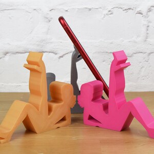 May include: Three phone stands made of plastic in the shape of people. One is orange, one is pink, and one is gray. The stands are designed to hold a phone upright.