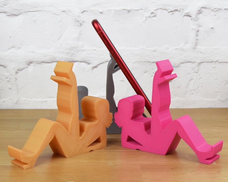 May include: Three phone stands made of plastic in the shape of people. One is orange, one is pink, and one is gray. The stands are designed to hold a phone upright.