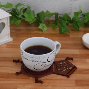 Puede incluir: Un posavasos hexagonal de cer&aacute;mica marr&oacute;n con detalles dorados y la palabra "Coffee" inscrita en el centro. Una taza de caf&eacute; blanca con letras negras que dice "smooth and rich cappuccino" se encuentra encima del posavasos.
