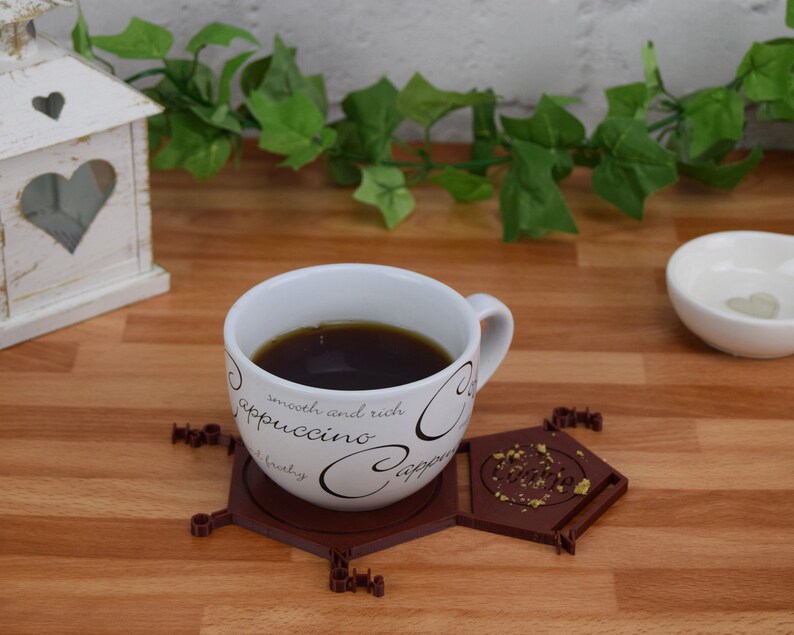 Puede incluir: Un posavasos hexagonal de cer&aacute;mica marr&oacute;n con detalles dorados y la palabra "Coffee" inscrita en el centro. Una taza de caf&eacute; blanca con letras negras que dice "smooth and rich cappuccino" se encuentra encima del posavasos.
