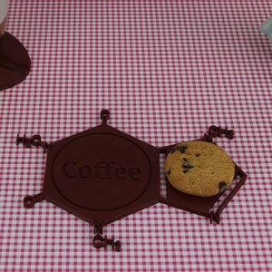 Puede incluir: Un posavasos hexagonal marr&oacute;n con la palabra "Coffee" inscrita en &eacute;l. Una galleta con chispas de chocolate descansa sobre el posavasos.
