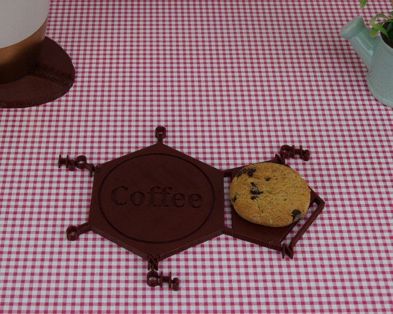 Puede incluir: Un posavasos hexagonal marr&oacute;n con la palabra "Coffee" inscrita en &eacute;l. Una galleta con chispas de chocolate descansa sobre el posavasos.