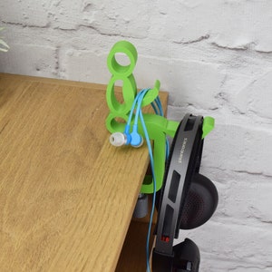 May include: Green 3D printed headphone and earbud holder shaped like a person sitting down. The holder is attached to a wooden desk. The headphones are black with the word "Plantronics" printed on the side.