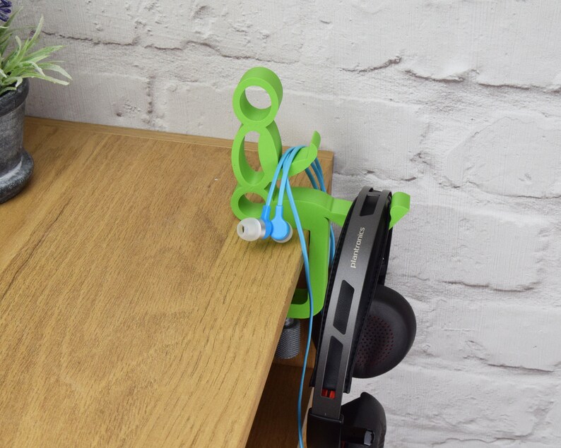 May include: Green 3D printed headphone and earbud holder shaped like a person sitting down. The holder is attached to a wooden desk. The headphones are black with the word "Plantronics" printed on the side.