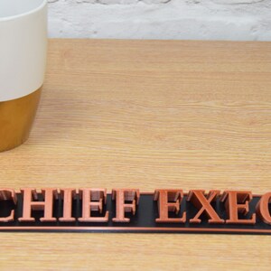 May include: A copper-colored desk nameplate with the words "CHIEF EXEC" on a black background.