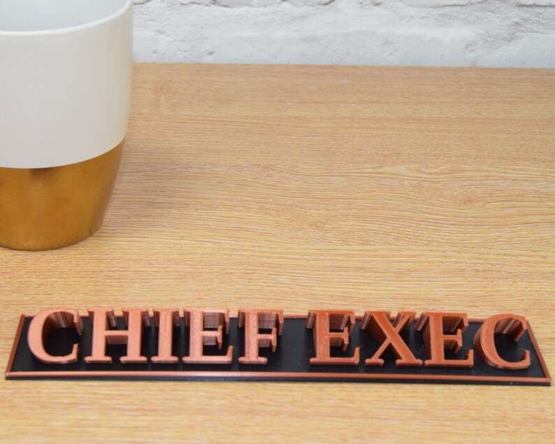 May include: A copper-colored desk nameplate with the words "CHIEF EXEC" on a black background.