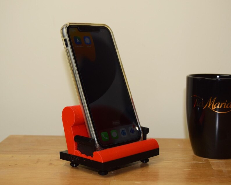 May include: A red and black phone stand with a smartphone in a clear case. The phone is displaying several apps on the screen. The stand is on a wooden surface.