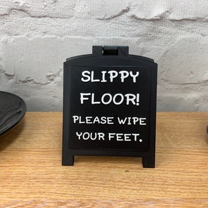 May include: A black, freestanding sign with the text "SLIPPY FLOOR! PLEASE WIPE YOUR FEET." The sign is on a wooden surface. The background includes a white brick wall, a coffee cup, and a small potted plant.