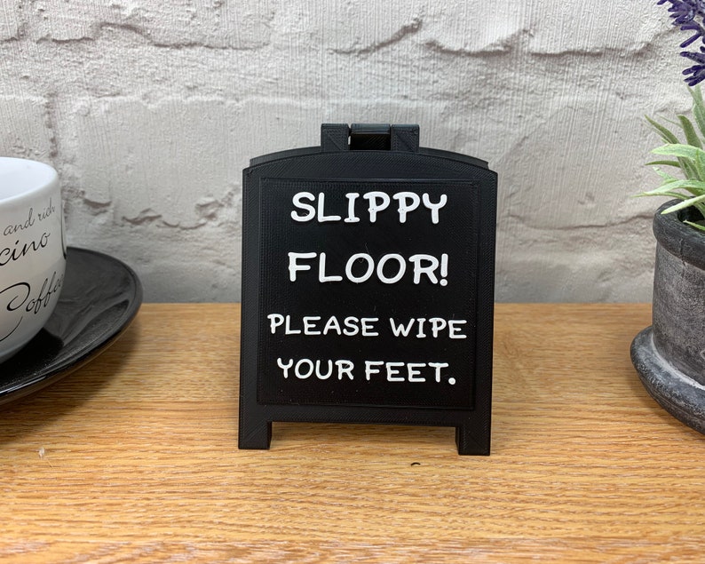 May include: A black, freestanding sign with the text "SLIPPY FLOOR! PLEASE WIPE YOUR FEET." The sign is on a wooden surface. The background includes a white brick wall, a coffee cup, and a small potted plant.