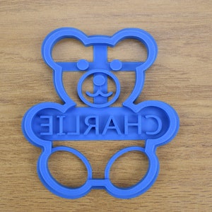 Puede incluir: Cortador de galletas de pl&aacute;stico azul con forma de osito de peluche con el nombre "CHARLIE" escrito en la parte delantera.