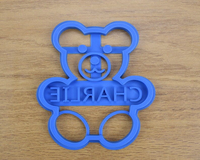 Puede incluir: Cortador de galletas de pl&aacute;stico azul con forma de osito de peluche con el nombre "CHARLIE" escrito en la parte delantera.