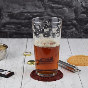 May include: A glass of beer with a foamy head in a clear pint glass with markings for half pint and pint. The glass is on a brown coaster with the word "Here" printed on it. A brown coaster with the text "Your words here" is on a white wooden surface.