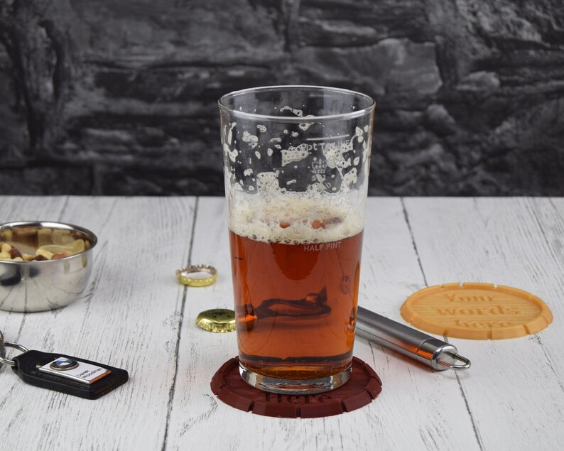 May include: A glass of beer with a foamy head in a clear pint glass with markings for half pint and pint. The glass is on a brown coaster with the word "Here" printed on it. A brown coaster with the text "Your words here" is on a white wooden surface.
