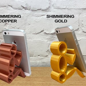 May include: Two 3D printed phone stands in the shape of a person sitting down. The stand on the left is a shimmering copper color and the stand on the right is a shimmering gold color.
