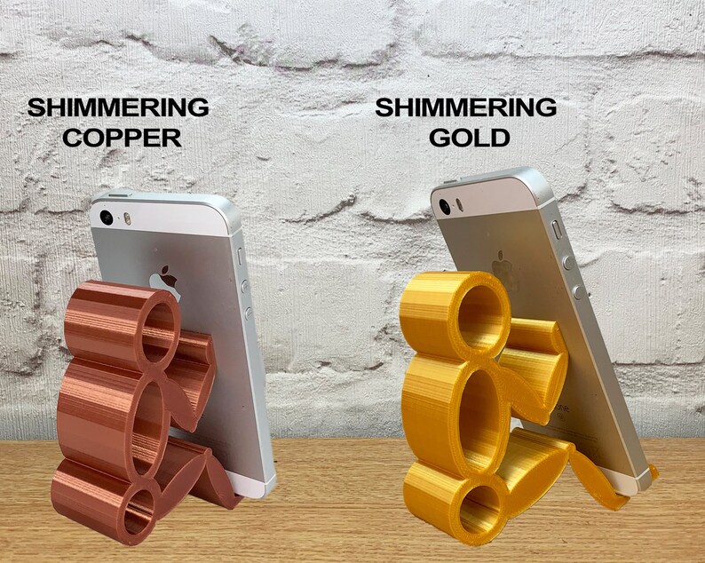 May include: Two 3D printed phone stands in the shape of a person sitting down. The stand on the left is a shimmering copper color and the stand on the right is a shimmering gold color.