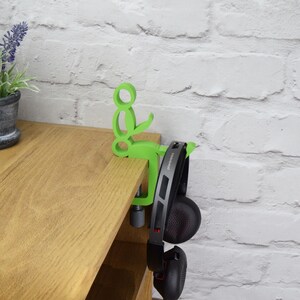 May include: A green plastic headphone holder attached to a wooden shelf. The holder is shaped like a person sitting down with their arms outstretched. A pair of black headphones are hanging from the holder.