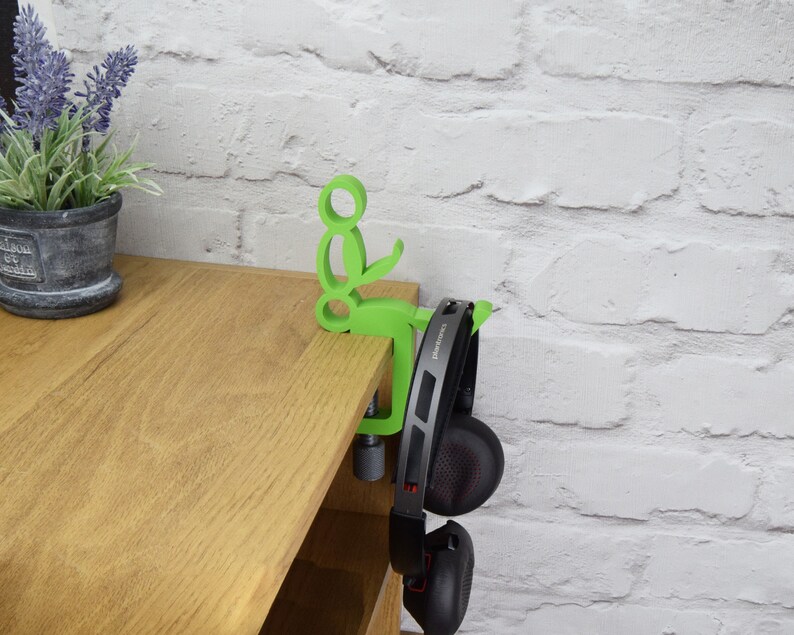May include: A green plastic headphone holder attached to a wooden shelf. The holder is shaped like a person sitting down with their arms outstretched. A pair of black headphones are hanging from the holder.