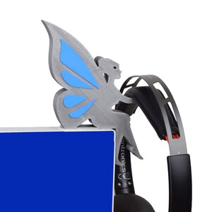 K&ouml;nnte beinhalten: Eine graue, 3D-gedruckte Feenfigur mit blauen Fl&uuml;geln, die auf einer blauen Oberfl&auml;che sitzt. Die Fee h&auml;lt ein schwarzes und rotes Headset.