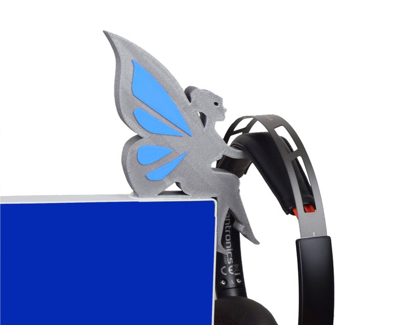 K&ouml;nnte beinhalten: Eine graue, 3D-gedruckte Feenfigur mit blauen Fl&uuml;geln, die auf einer blauen Oberfl&auml;che sitzt. Die Fee h&auml;lt ein schwarzes und rotes Headset.