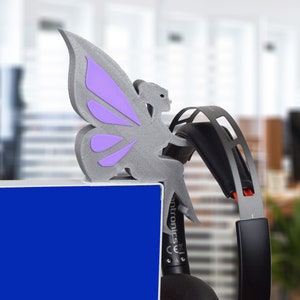 K&ouml;nnte beinhalten: Eine graue, im 3D-Druck hergestellte Feenfigur mit lila Fl&uuml;geln, die auf einer blauen Oberfl&auml;che mit einem schwarzen und roten Headset sitzt.