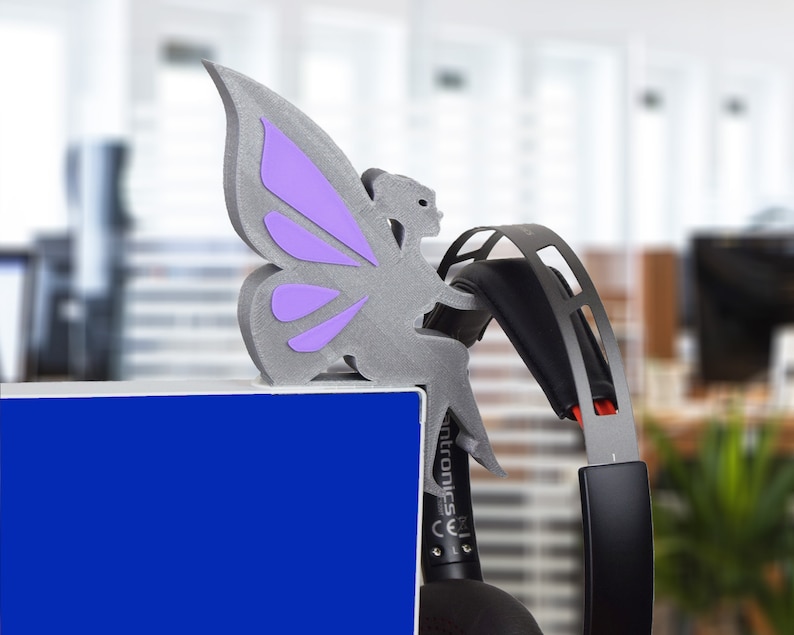 K&ouml;nnte beinhalten: Eine graue, im 3D-Druck hergestellte Feenfigur mit lila Fl&uuml;geln, die auf einer blauen Oberfl&auml;che mit einem schwarzen und roten Headset sitzt.