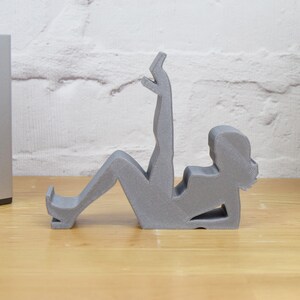 May include: A gray 3D printed figurine of a person sitting with their legs raised. The figurine is designed to hold a phone or other small object.