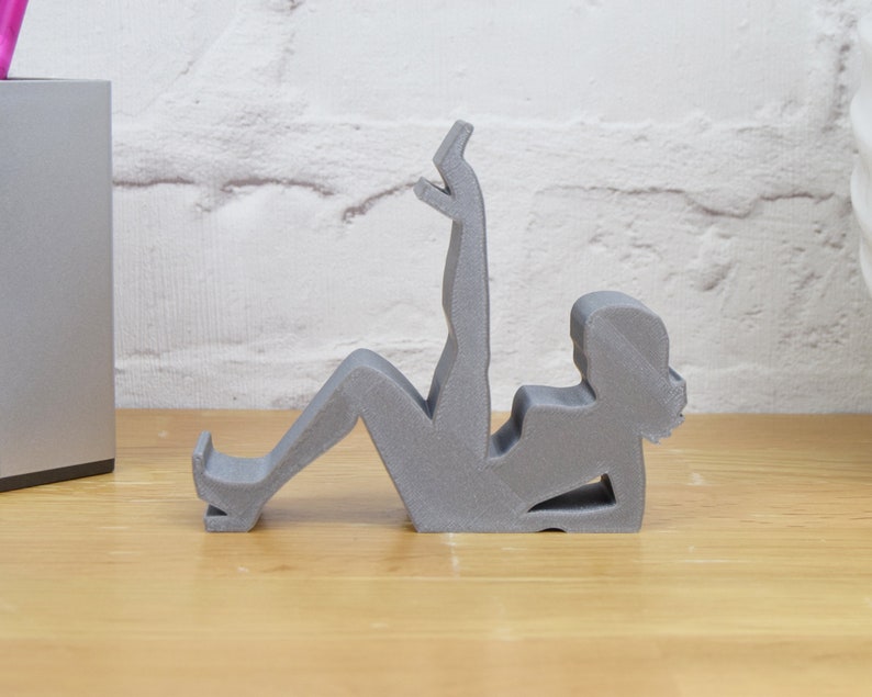 May include: A gray 3D printed figurine of a person sitting with their legs raised. The figurine is designed to hold a phone or other small object.