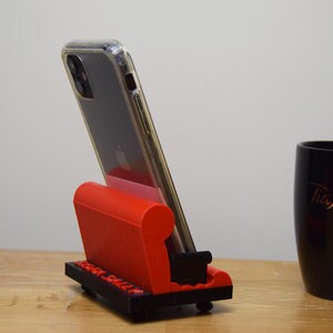 May include: A red and black 3D printed phone stand shaped like a couch. A black smartphone with a clear case is in the stand.