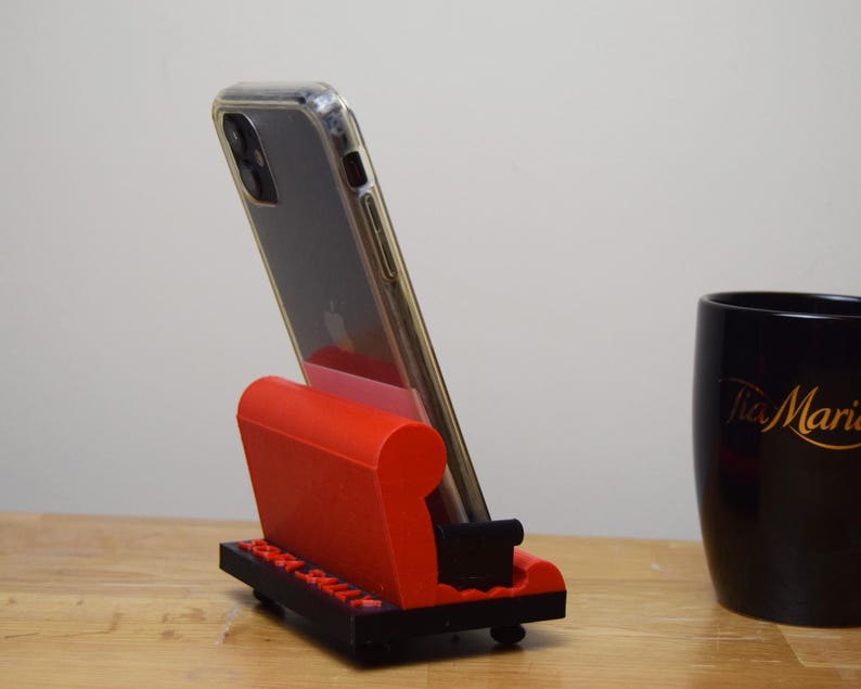 May include: A red and black 3D printed phone stand shaped like a couch. A black smartphone with a clear case is in the stand.