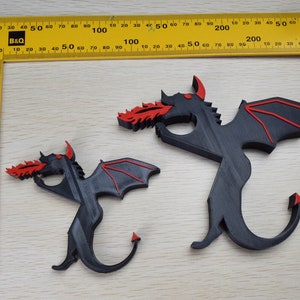 May include: Two black dragon figurines with red accents, one larger than the other, are displayed on a light wood surface. The dragons have outstretched wings and are spewing red flames. A yellow ruler with black markings is in the background.