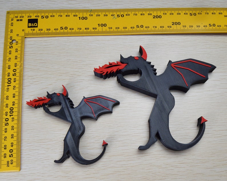 May include: Two black dragon figurines with red accents, one larger than the other, are displayed on a light wood surface. The dragons have outstretched wings and are spewing red flames. A yellow ruler with black markings is in the background.