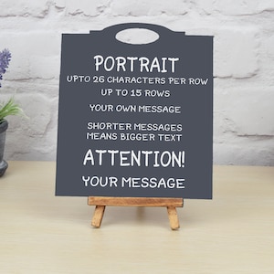 May include: A dark gray chalkboard easel with a white frame and a white handle. The chalkboard has the text "PORTRAIT", "UPTO 26 CHARACTERS PER ROW", "UP TO 15 ROWS", "YOUR OWN MESSAGE", "SHORTER MESSAGES MEANS BIGGER TEXT", "ATTENTION!" and "YOUR MESSAGE".