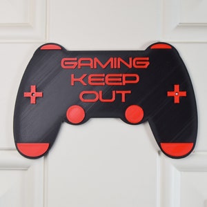 以下が含まれることがあります： 「GAMING KEEP OUT」というテキストが書かれた、ビデオゲームコントローラーの形をした黒と赤の3Dプリントサイン。