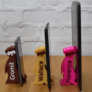 May include: Three 3D printed phone stands, each featuring a different color and the name of the stand printed on the front. The stands are holding up three different sized phones, from smallest to largest. The stands are brown, yellow, and pink.