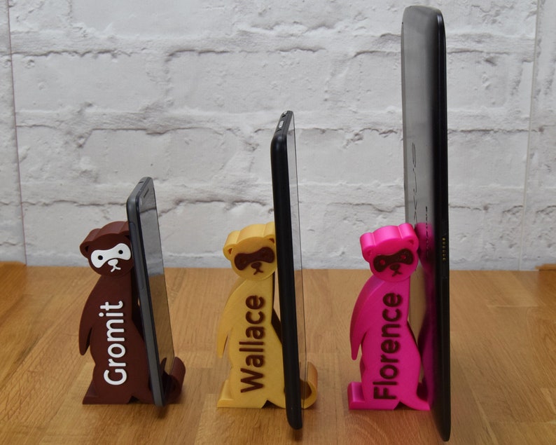 May include: Three 3D printed phone stands, each featuring a different color and the name of the stand printed on the front. The stands are holding up three different sized phones, from smallest to largest. The stands are brown, yellow, and pink.
