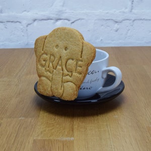 May include: A light brown cookie shaped like an elephant with the word "GRACE" embossed on it. The cookie is sitting on a black saucer next to a white coffee cup.