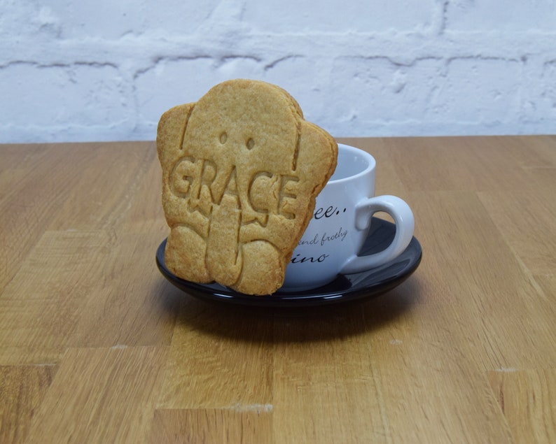May include: A light brown cookie shaped like an elephant with the word "GRACE" embossed on it. The cookie is sitting on a black saucer next to a white coffee cup.