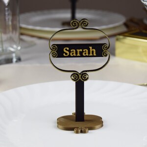 May include: A black and gold table place card with the name "Sarah" written in gold lettering.