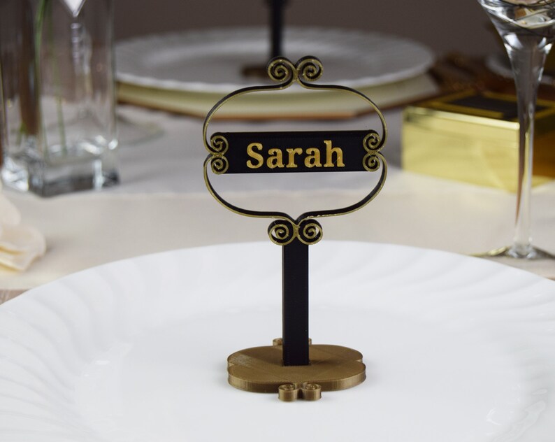 May include: A black and gold table place card with the name "Sarah" written in gold lettering.
