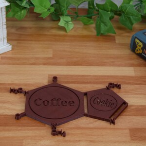 Puede incluir: Un posavasos hexagonal marr&oacute;n con las palabras "Coffee" y "Cookie" impresas en &eacute;l. El posavasos est&aacute; hecho de un material impreso en 3D y tiene un dise&ntilde;o geom&eacute;trico.