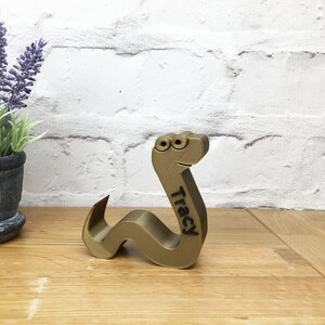 May include: A gold-colored, 3D printed worm with googly eyes and the name "Tracy" inscribed on its side. The worm is sitting on a wooden surface.