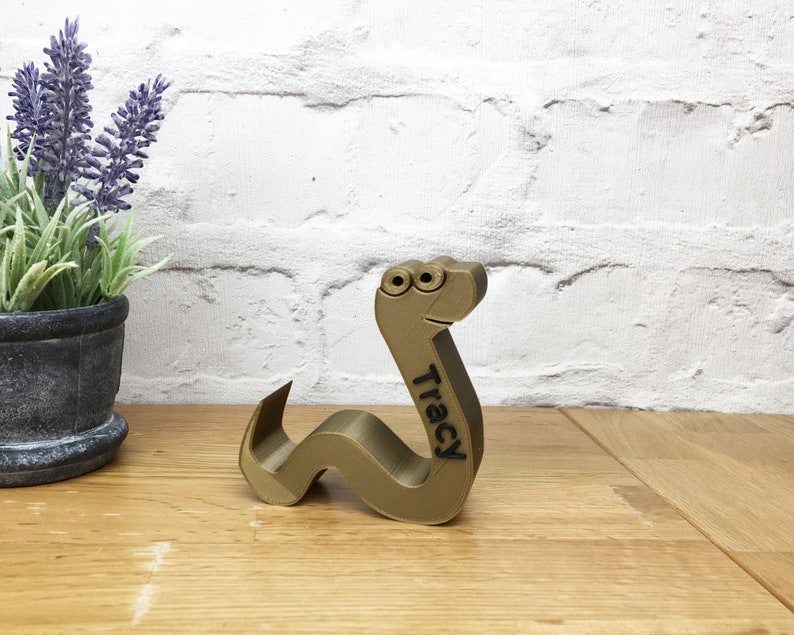 May include: A gold-colored, 3D printed worm with googly eyes and the name "Tracy" inscribed on its side. The worm is sitting on a wooden surface.