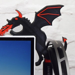 May include: Black and red dragon-shaped headset holder. The dragon is perched on the edge of a white monitor, with its wings and flames in red. The holder is designed to hold a headset, which is visible in the image.