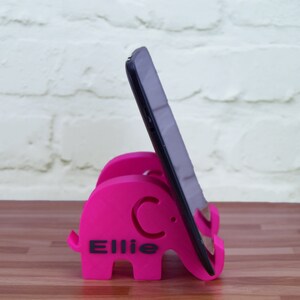 Pode incluir: Um suporte de telefone cor-de-rosa em forma de elefante com o nome "Ellie" impresso nele. Um tablet preto est&aacute; apoiado no suporte.