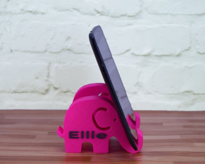 Pode incluir: Um suporte de telefone cor-de-rosa em forma de elefante com o nome "Ellie" impresso nele. Um tablet preto est&aacute; apoiado no suporte.